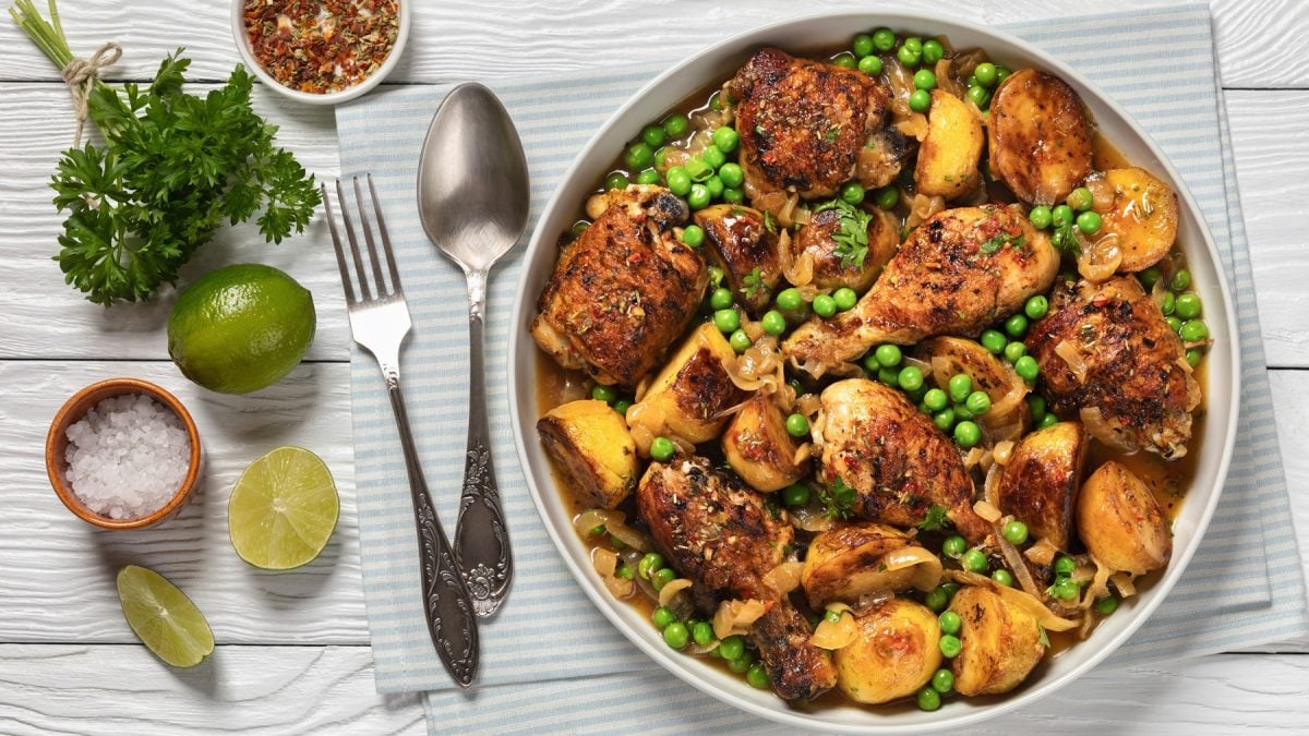 Saftig gebratenes Hähnchen mit grünen Erbsen, gelben Kartoffeln und Zwiebelringen in Schale, daneben Limetten, Salz und Besteck.