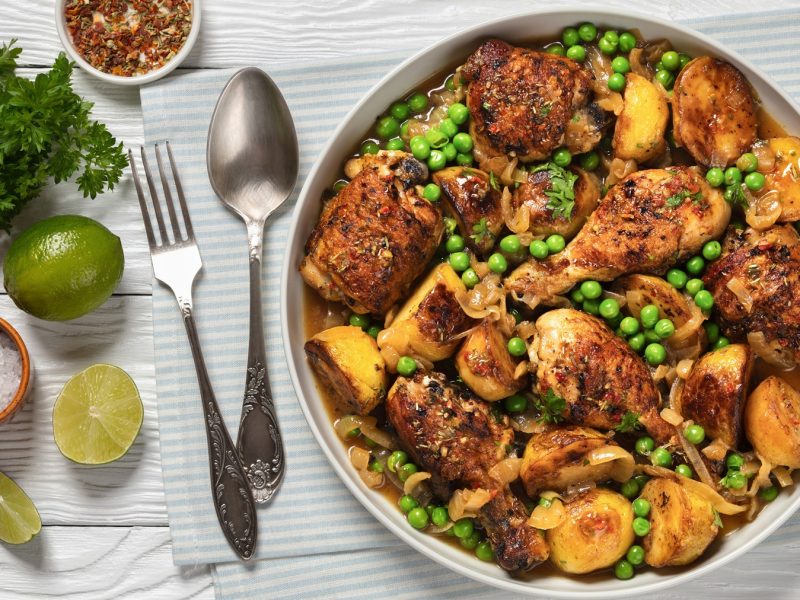 Saftig gebratenes Hähnchen mit grünen Erbsen, gelben Kartoffeln und Zwiebelringen in Schale, daneben Limetten, Salz und Besteck.