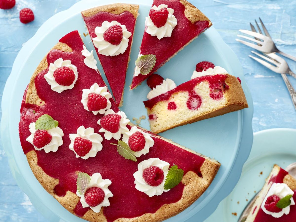 Express-Himbeerkuchen auf einem blauen Teller vor blauem Hintergrund. Rechts davon Besteck, links ein Schälchen mit Himbeeren.