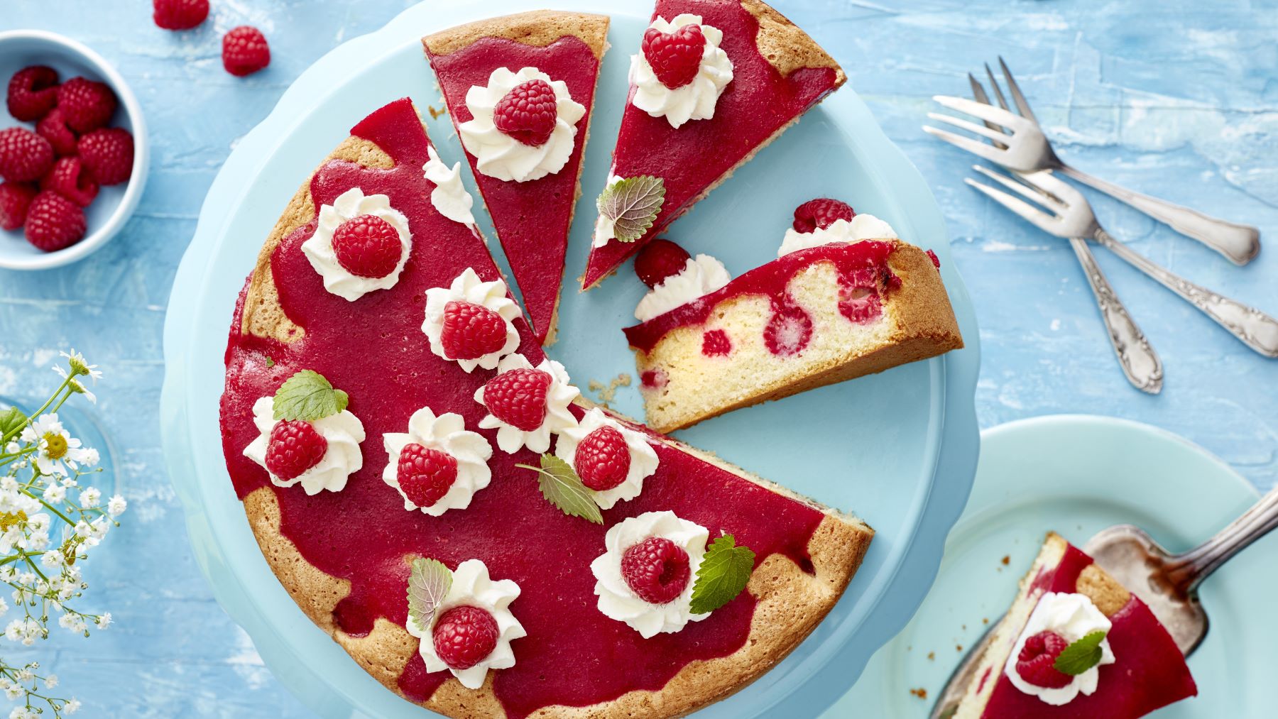 Express-Himbeerkuchen auf einem blauen Teller vor blauem Hintergrund. Rechts davon Besteck, links ein Schälchen mit Himbeeren.