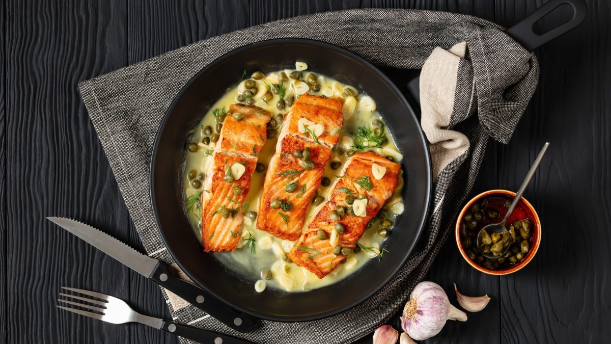 Nahaufnahme von gebratenem Lachs in heller Sauce mit Dill, Knoblauch und Kapern, daneben Besteck und frischer Knoblauch.
