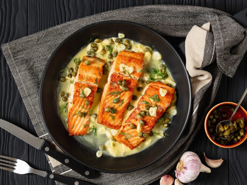 Nahaufnahme von gebratenem Lachs in heller Sauce mit Dill, Knoblauch und Kapern, daneben Besteck und frischer Knoblauch.