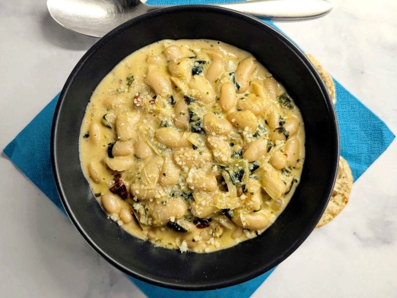Schwarzer Teller auf weißem Marmor. Darauf cremige Marry Me Beans in cremiger Sauce.