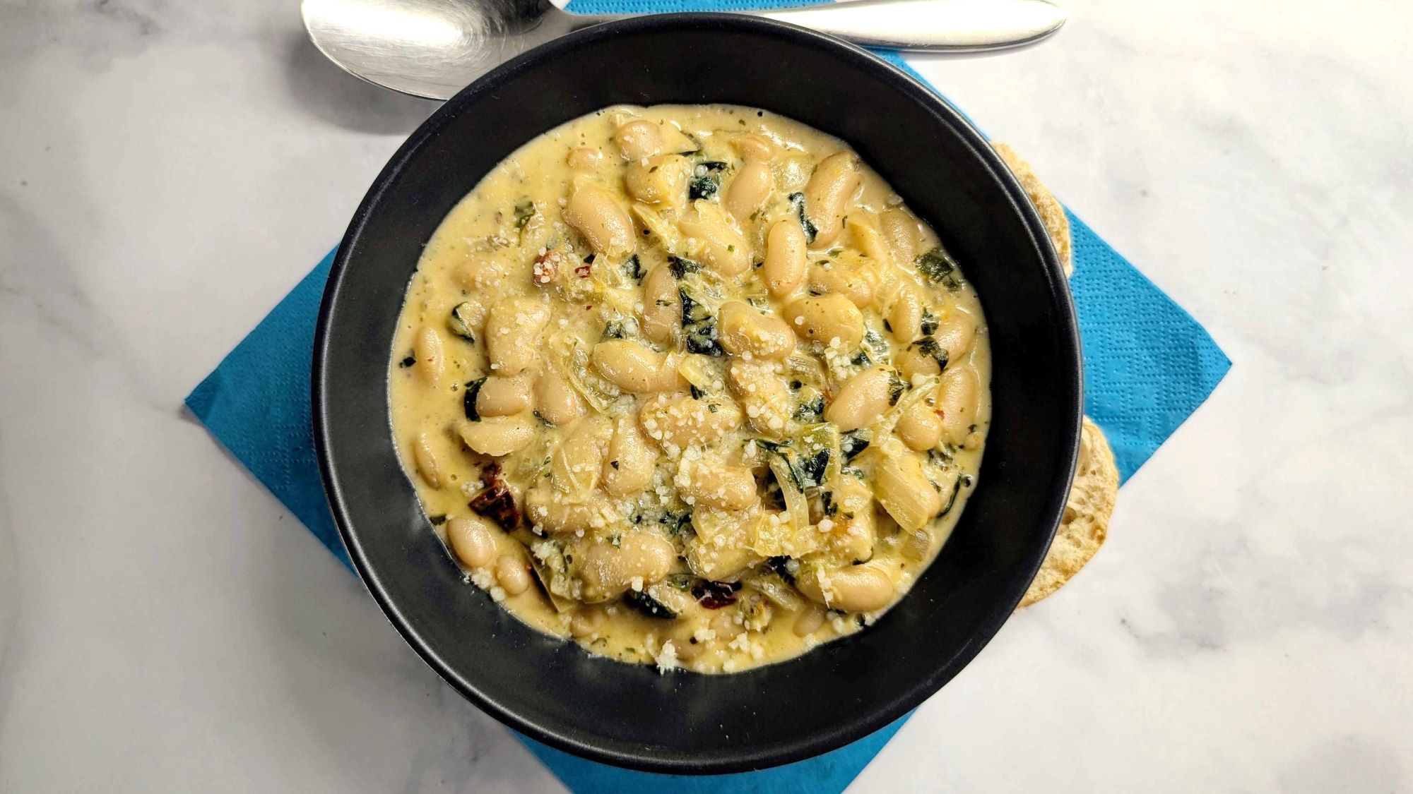 Schwarzer Teller auf weißem Marmor. Darauf cremige Marry Me Beans in cremiger Sauce.