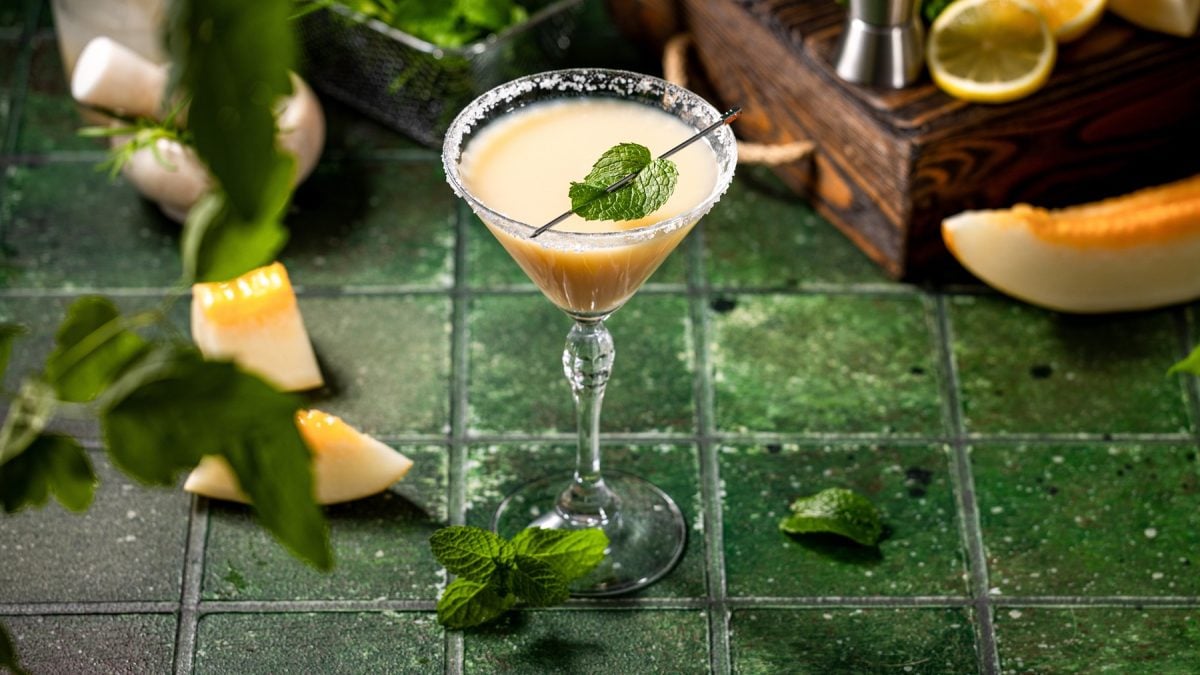 Gelblich-beiger Margarita mit Minzdeko in Glas mit Salzrand, arrangiert auf rustikalem grünem Untergrund mit Melone und Kräutern.
