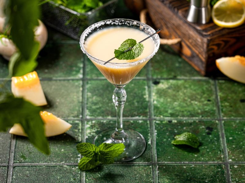 Gelblich-beiger Margarita mit Minzdeko in Glas mit Salzrand, arrangiert auf rustikalem grünem Untergrund mit Melone und Kräutern.