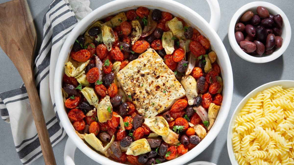 Mediterraner Feta-Auflauf mit goldbraunem Käse, gerösteten Tomaten, Oliven und Artischocken, daneben gekochte Fusilli-Nudeln.