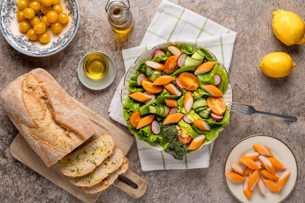 Frischer Sommersalat mit Surimi, Gurken, Radieschen und gelben Tomaten auf weißem Tuch, daneben Brot, Öl und Zitrusfrüchte.
