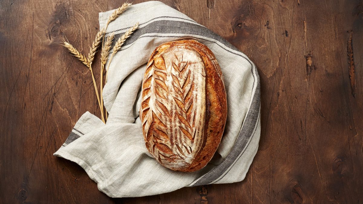 Rustikales Sauerteigbrot mit kunstvoll eingeritztem Muster liegt auf einem Leinentuch neben Ähren auf dunklem Holztisch.