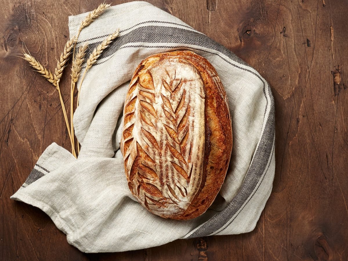 Rustikales Sauerteigbrot mit kunstvoll eingeritztem Muster liegt auf einem Leinentuch neben Ähren auf dunklem Holztisch.