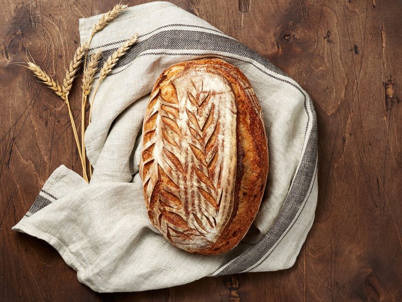 Rustikales Sauerteigbrot mit kunstvoll eingeritztem Muster liegt auf einem Leinentuch neben Ähren auf dunklem Holztisch.
