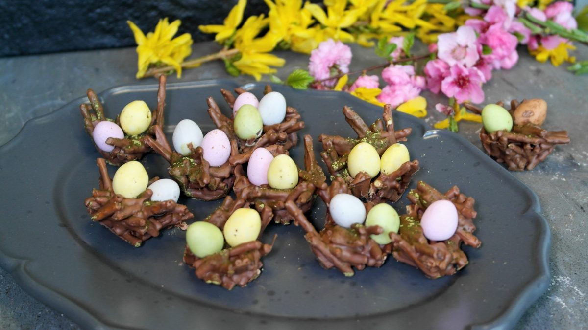 Ein grauer Teller mit den Schokonestern und kleinen Schokoeiern auf einem grauen Steintisch mit schwarzem Hintergrund und gelben sowie pinken Blüten.