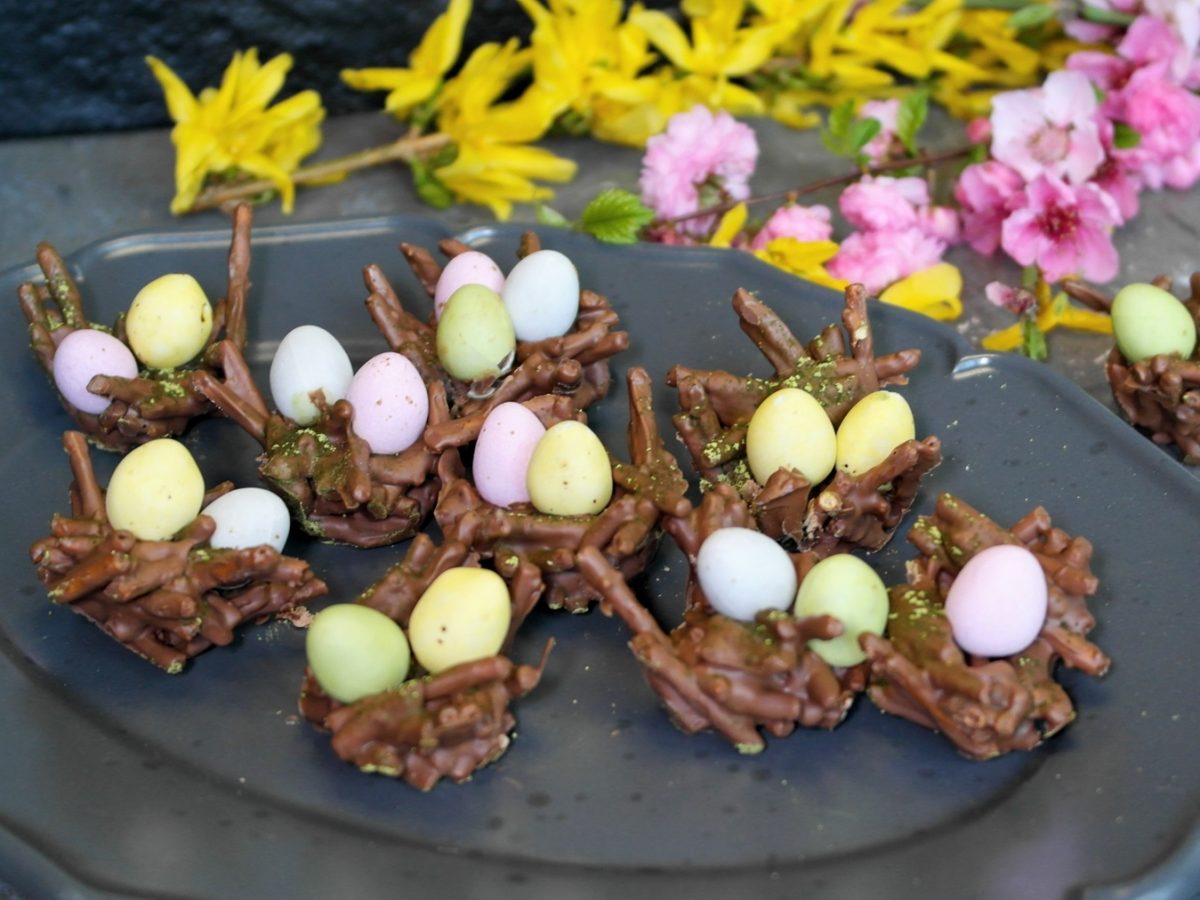 Ein grauer Teller mit den Schokonestern und kleinen Schokoeiern auf einem grauen Steintisch mit schwarzem Hintergrund und gelben sowie pinken Blüten.