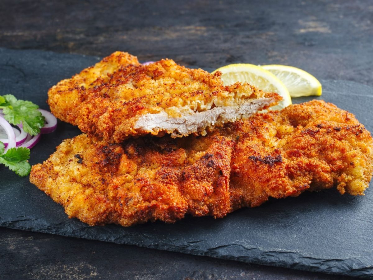 Knusprig paniertes Schnitzel auf schwarzer Steinplatte mit roten Zwiebelringen, Petersilie und Zitronenscheiben.