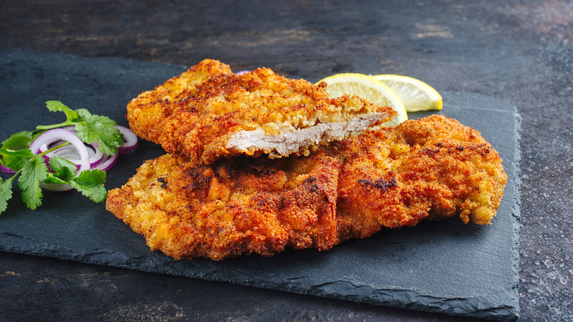 Knusprig paniertes Schnitzel auf schwarzer Steinplatte mit roten Zwiebelringen, Petersilie und Zitronenscheiben.