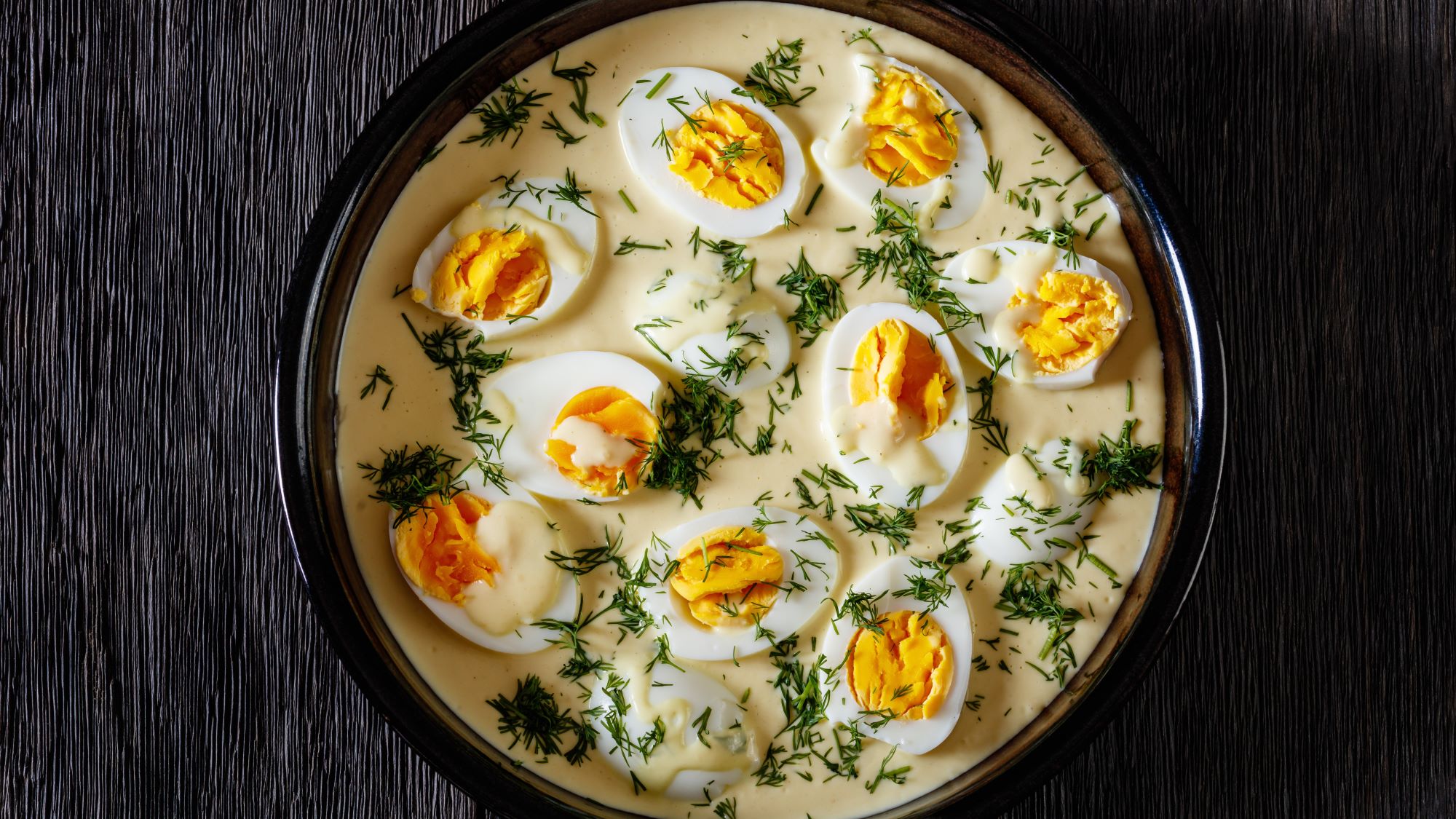 Hart gekochte Eier in cremiger Sauce mit Kräuterdekor, angerichtet in einer schwarzen Bowl mit mattem Finish.