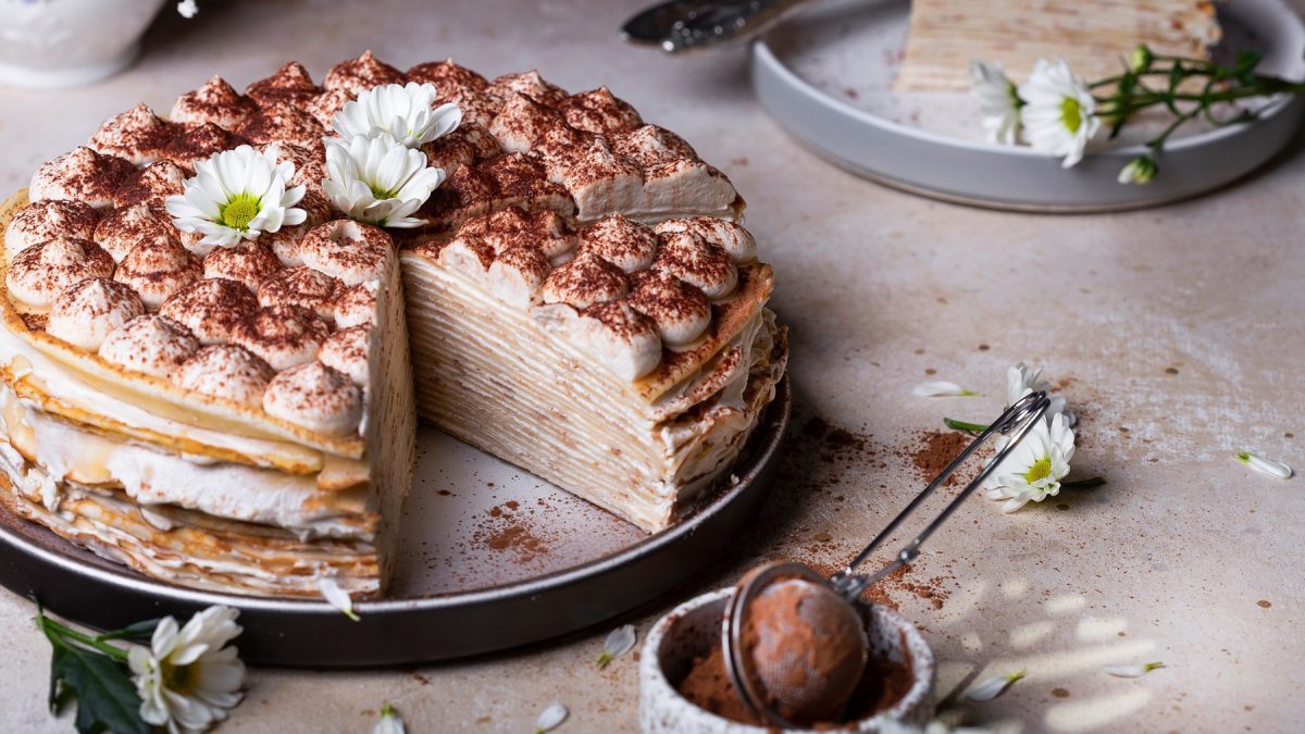 Aufgeschnittene Tiramisu-Crêpe-Torte mit Kakaopulver und Sahnetupfen, dekoriert mit zwei weißen Chrysanthemen auf hellem Untergrund.