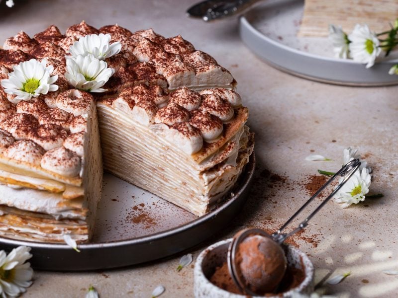 Aufgeschnittene Tiramisu-Crêpe-Torte mit Kakaopulver und Sahnetupfen, dekoriert mit zwei weißen Chrysanthemen auf hellem Untergrund.