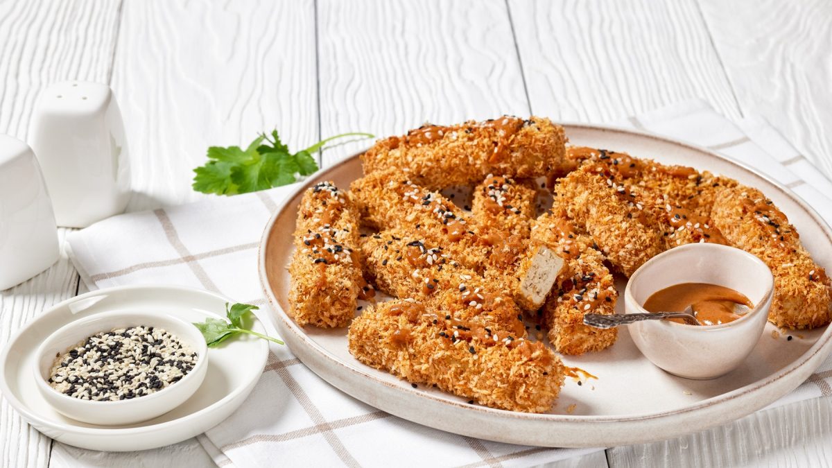 Knusprige goldbraune Tofu-Sticks mit Sesam auf einem Teller, dazu cremiger Dip und Petersilie auf weißem Holztisch.