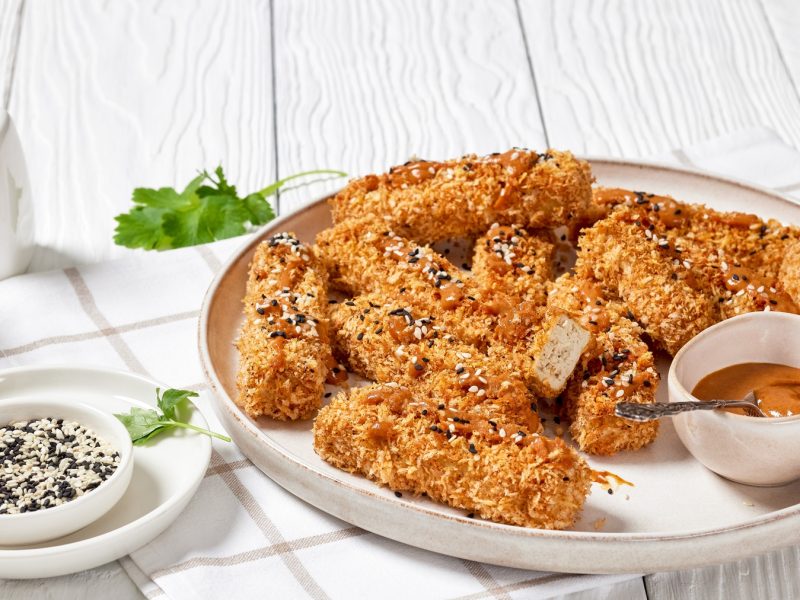 Knusprige goldbraune Tofu-Sticks mit Sesam auf einem Teller, dazu cremiger Dip und Petersilie auf weißem Holztisch.