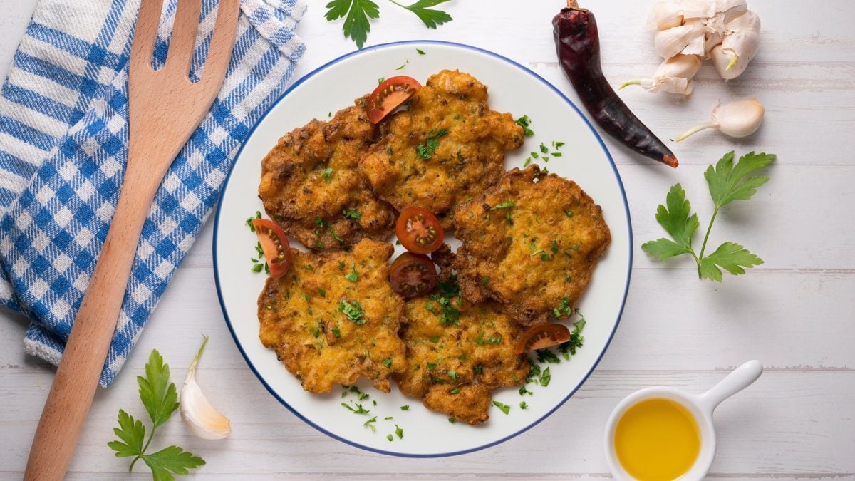 Sechs goldbraune Tortillitas de Camarones mit frischer Petersilie garniert, angerichtet auf hellem Holztisch mit Kräutern und Öl.
