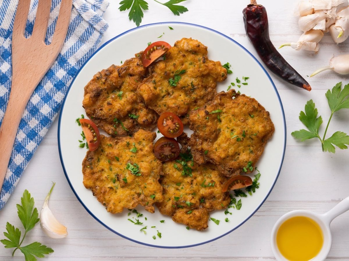 Sechs goldbraune Tortillitas de Camarones mit frischer Petersilie garniert, angerichtet auf hellem Holztisch mit Kräutern und Öl.