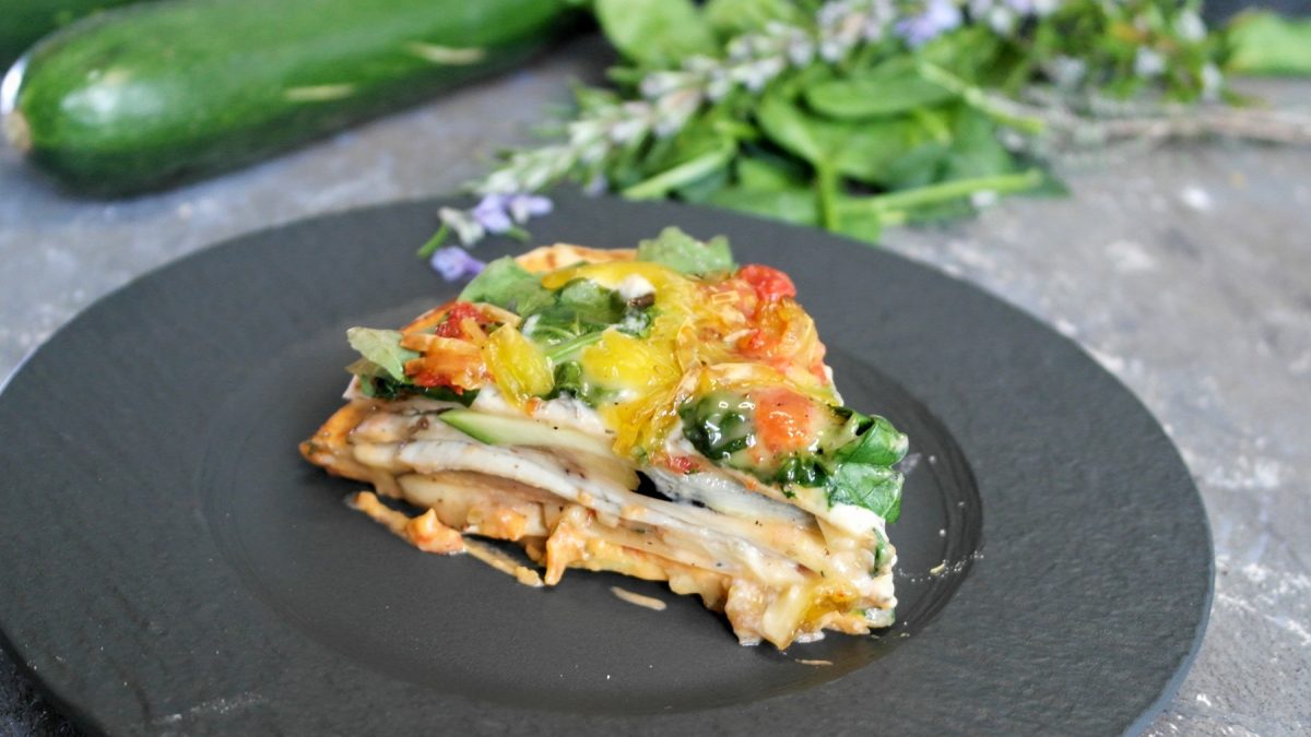 Ein schwarzer Teller mit einem Stück der Wrap-Lasagne auf grauem Untergrund, mit Kräutern und Zucchini im Hintergrund.