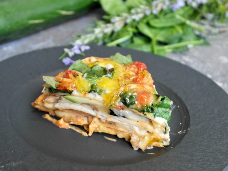 Ein schwarzer Teller mit einem Stück der Wrap-Lasagne auf grauem Untergrund, mit Kräutern und Zucchini im Hintergrund.