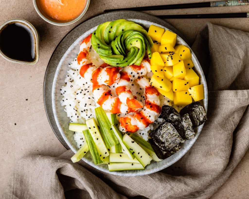 Bunte Poké Bowl mit Reis, Garnelen, Avocado-Rose, Mango, Gurkenstreifen und geröstetem Nori auf hellem Tuch.