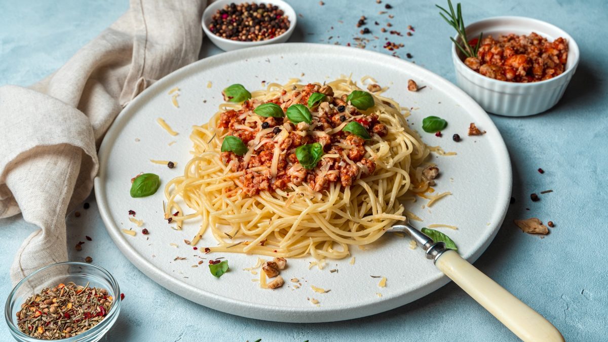 Ein weißer Teller mit den Spaghetti und der Walnuss-Pilz-Bolognese auf einem hellblauen Untergund, mit Gewürzen in weißen Schälchen darum platziert. Alles von schräg oben fotografiert.