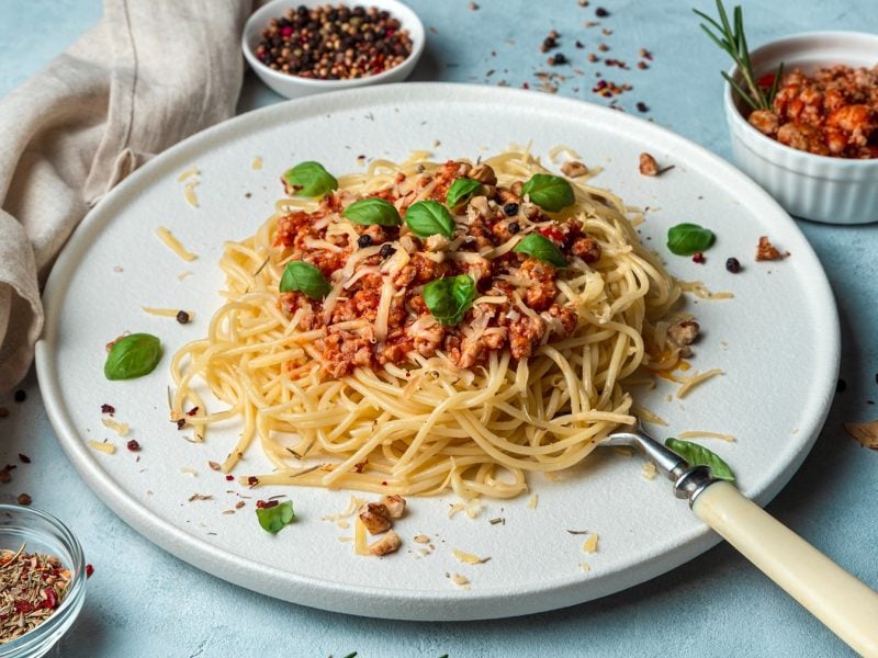 Ein weißer Teller mit den Spaghetti und der Walnuss-Pilz-Bolognese auf einem hellblauen Untergund, mit Gewürzen in weißen Schälchen darum platziert. Alles von schräg oben fotografiert.