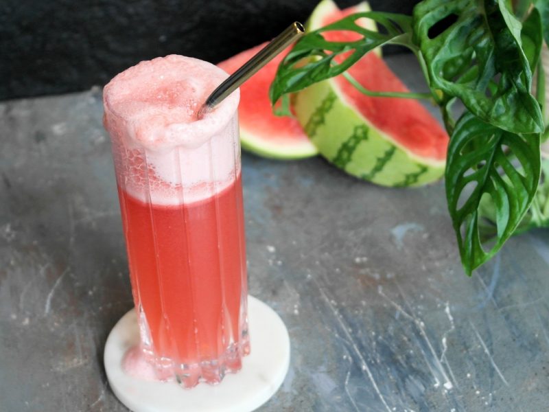 Ein Glas mit dem roten Cocktail auf einem grauen Untergrund. Alles von schräg oben fotografiert.