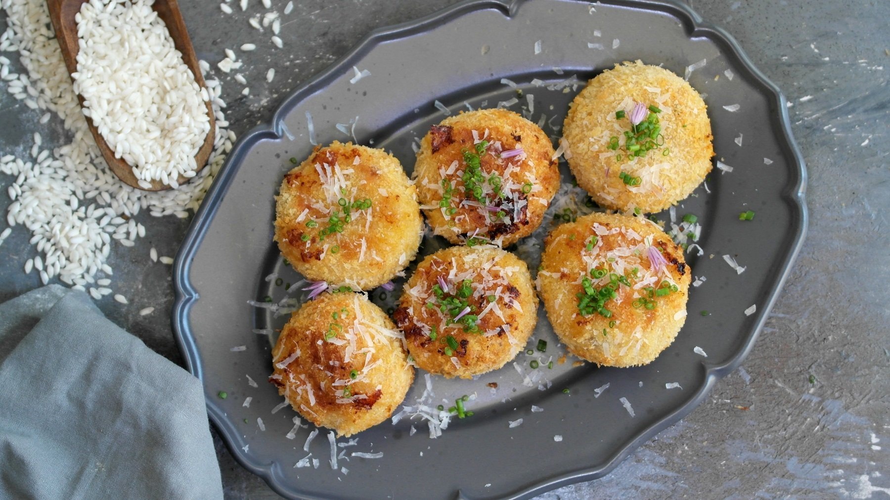 Ein grauer Teller mit den Arancini auf steinernem Untergrund. Daneben noch roher Risottoreis und ein graues Tuch. Alles direkt von oben fotografiert.