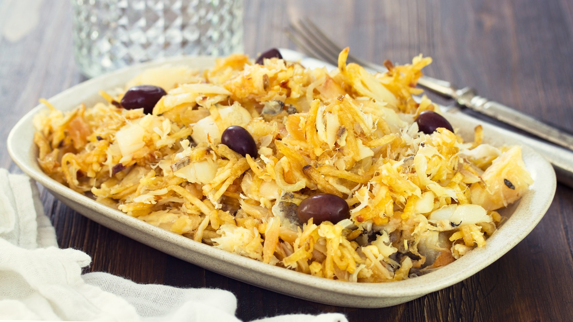 Blick auf einen weißen Teller mit Bacalhau à Brás, der auf einem grauen Holzuntergrund steht.