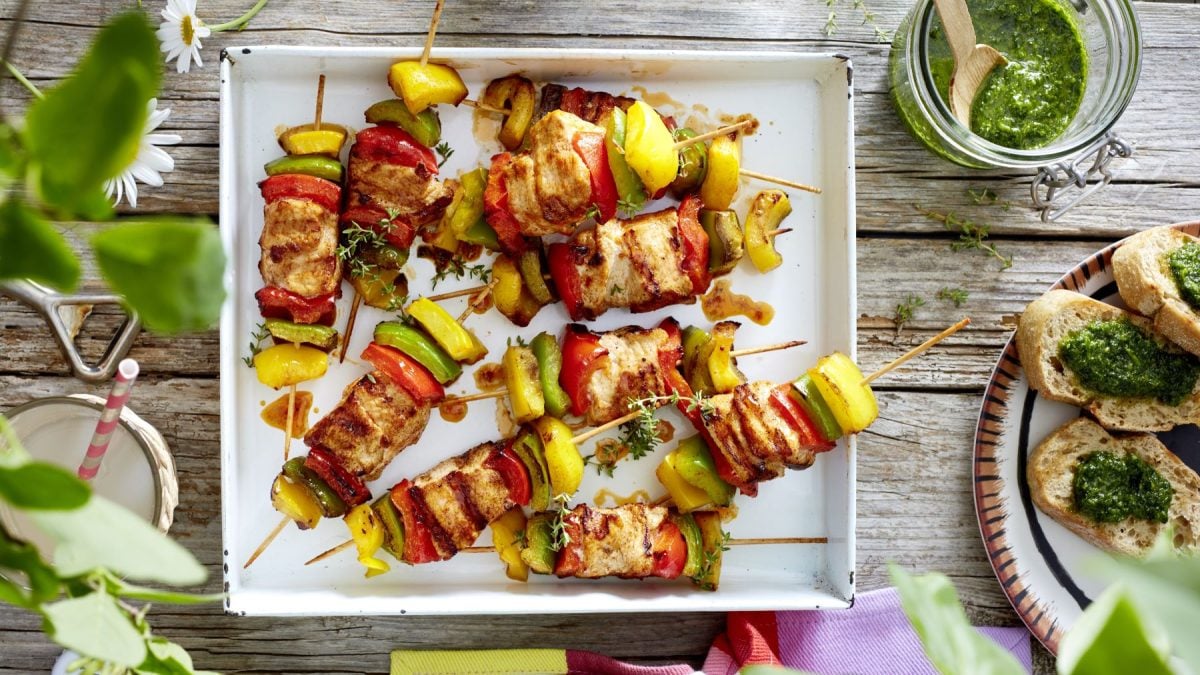 Paprika-Puten-Spieße auf einem weißen Teller vor hölzernem Hintergrund. Links auf dem Foto Pflanzen und rechts Pesto und Baguette.
