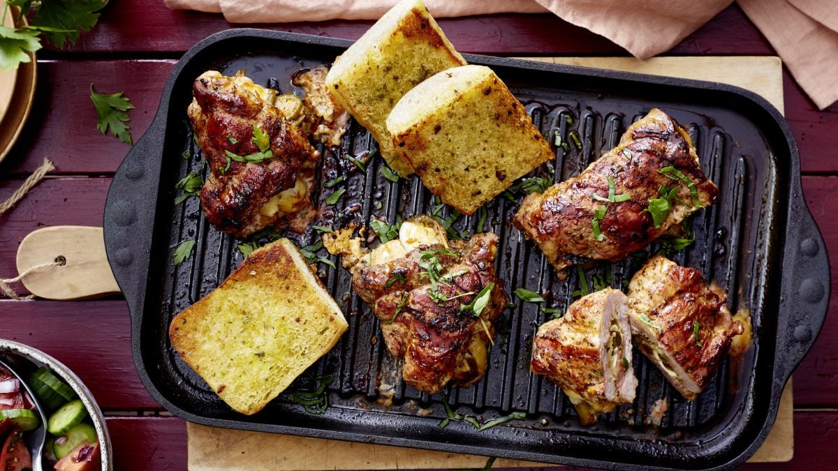 Zartes Cordon Bleu mit goldener Kruste auf Grillpfanne, flankiert von knusprig geröstetem Kräuterbrot auf rotem Holztisch.