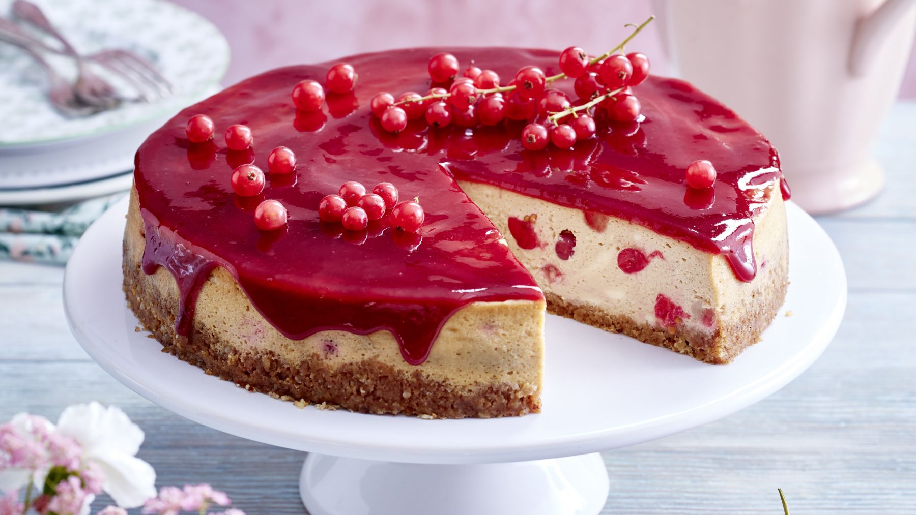 Tiramisu-Johannisbeer-Cheesecake auf einem weißen Tortenteller vor grauem Hintergrund. Hinten auf dem Foto Teller und Gabeln verteilt. Vorne links sind Blumen.