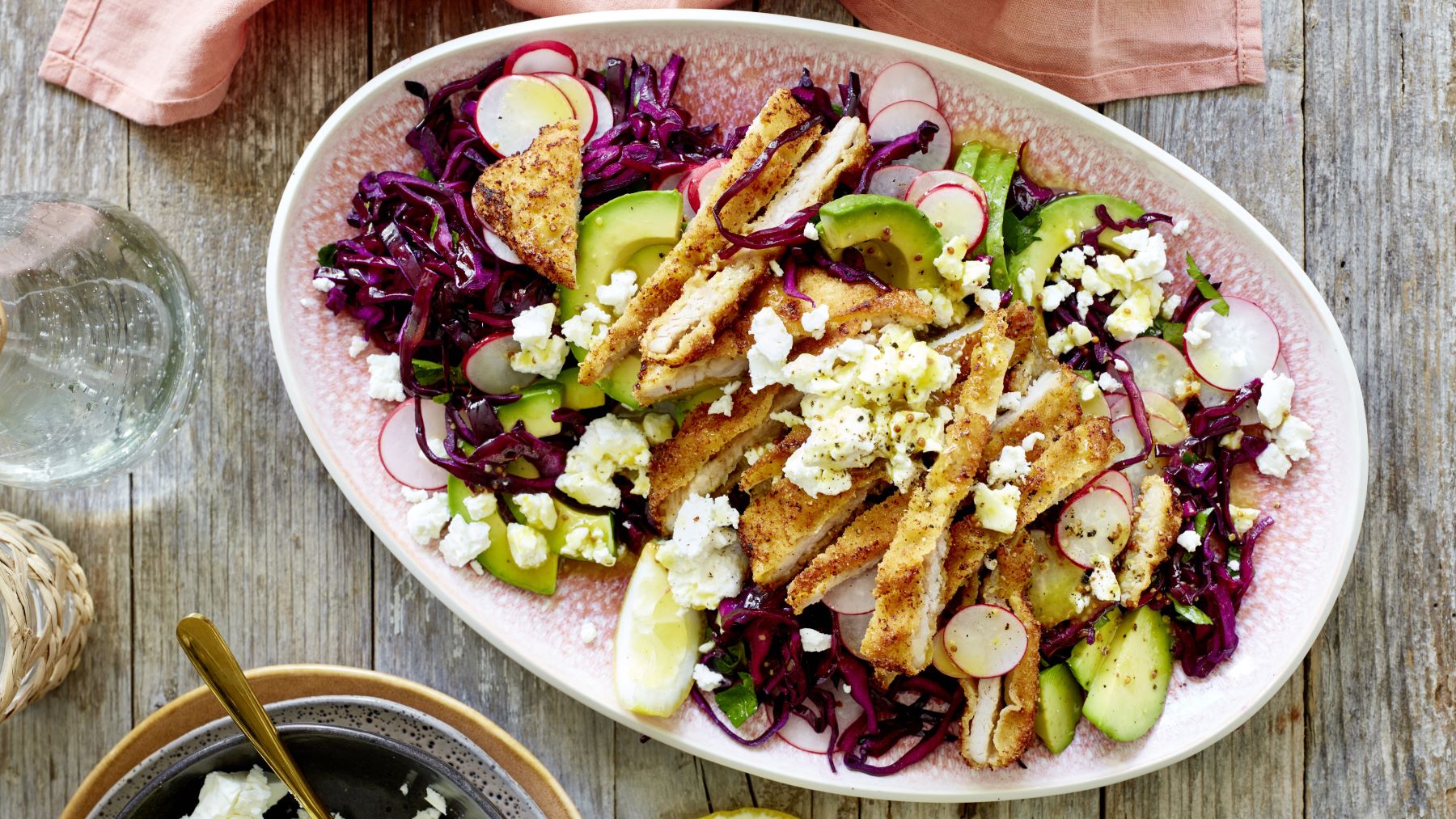Bunte Salatplatte mit Schnitzel auf einem roten Teller vor hölzernem Hintergrund. Oben auf dem Foto ein rosa Geschirrtuch und links ein Glas Wasser und Schüsselchen. Der Hintergrund ist hölzern.