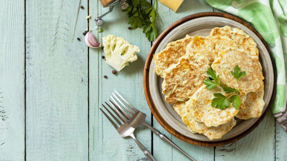 Stapel herzhafter Pfannkuchen mit Blumenkohl und Käse auf rustikalem Teller, flankiert von Gabeln, Kräutern und frischem Gemüse.