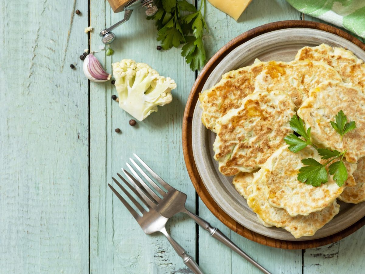 Stapel herzhafter Pfannkuchen mit Blumenkohl und Käse auf rustikalem Teller, flankiert von Gabeln, Kräutern und frischem Gemüse.