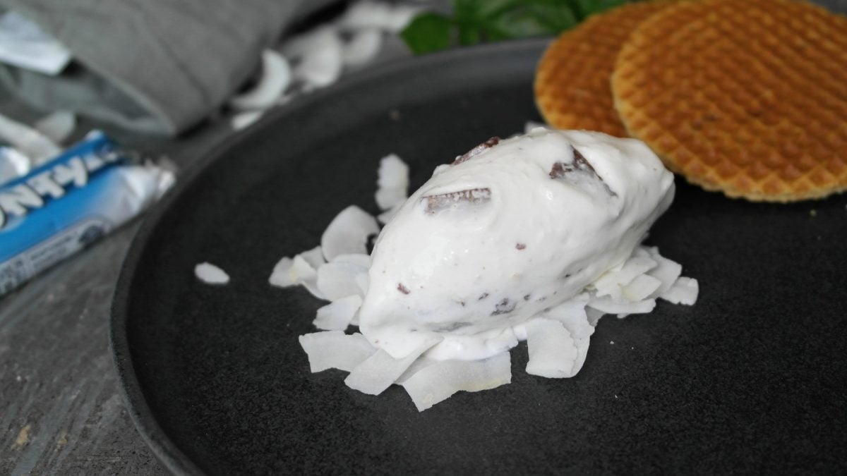 Eine Nocke der Eiscreme auf Kokoschips und einem dunklen Teller. Im Hintegrund Waffeln und ein Bounty Riegel erkennbar.