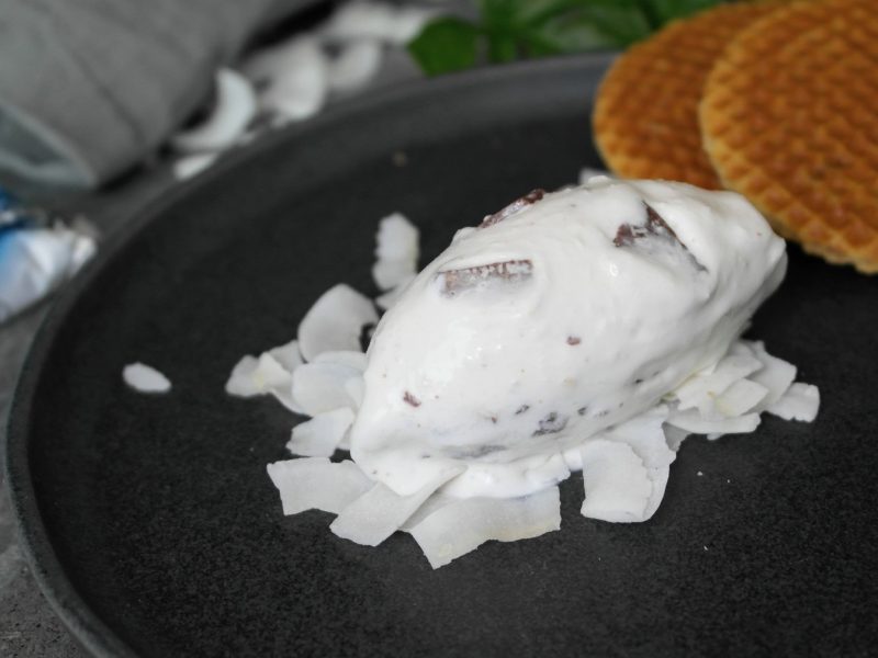 Eine Nocke der Eiscreme auf Kokoschips und einem dunklen Teller. Im Hintegrund Waffeln und ein Bounty Riegel erkennbar.
