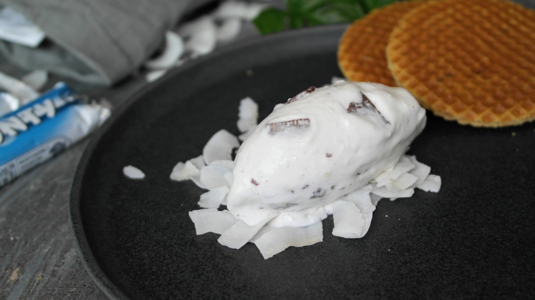 Eine Nocke der Eiscreme auf Kokoschips und einem dunklen Teller. Im Hintegrund Waffeln und ein Bounty Riegel erkennbar.