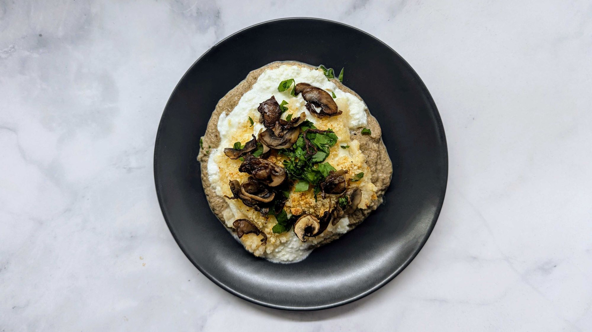 Burrata mit Champignons, frischer Petersilie und knusprigem Topping auf herzhafter Creme, serviert auf schwarzem Teller.