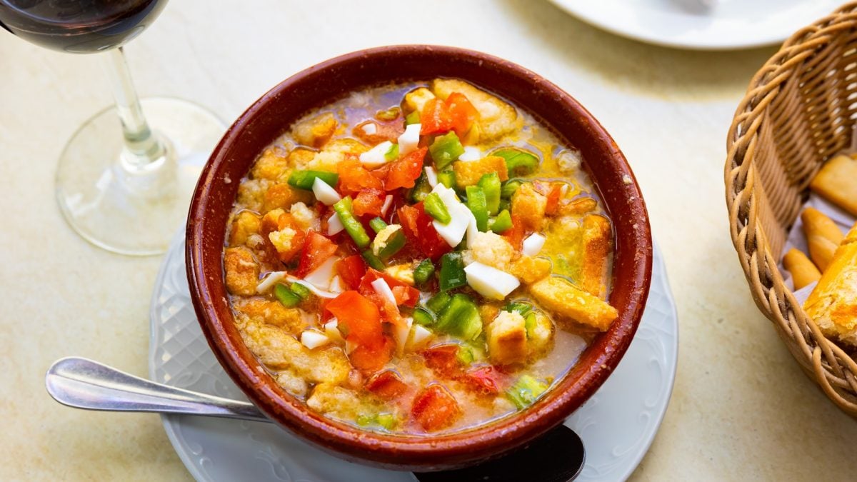 Spanischer Cojondongo mit Brotstücken, gehacktem Gemüse und Ei, serviert in brauner Schale neben Brotkorb und Weinglas.