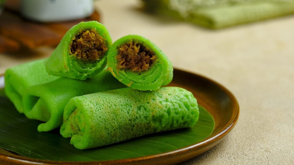 Die grünen Pfannkuchen-Röllchen mit Kokosfüllung auf einem schwarzen Teller und hellem Holztisch. Alles nah und von leicht schräg oben fotografiert.