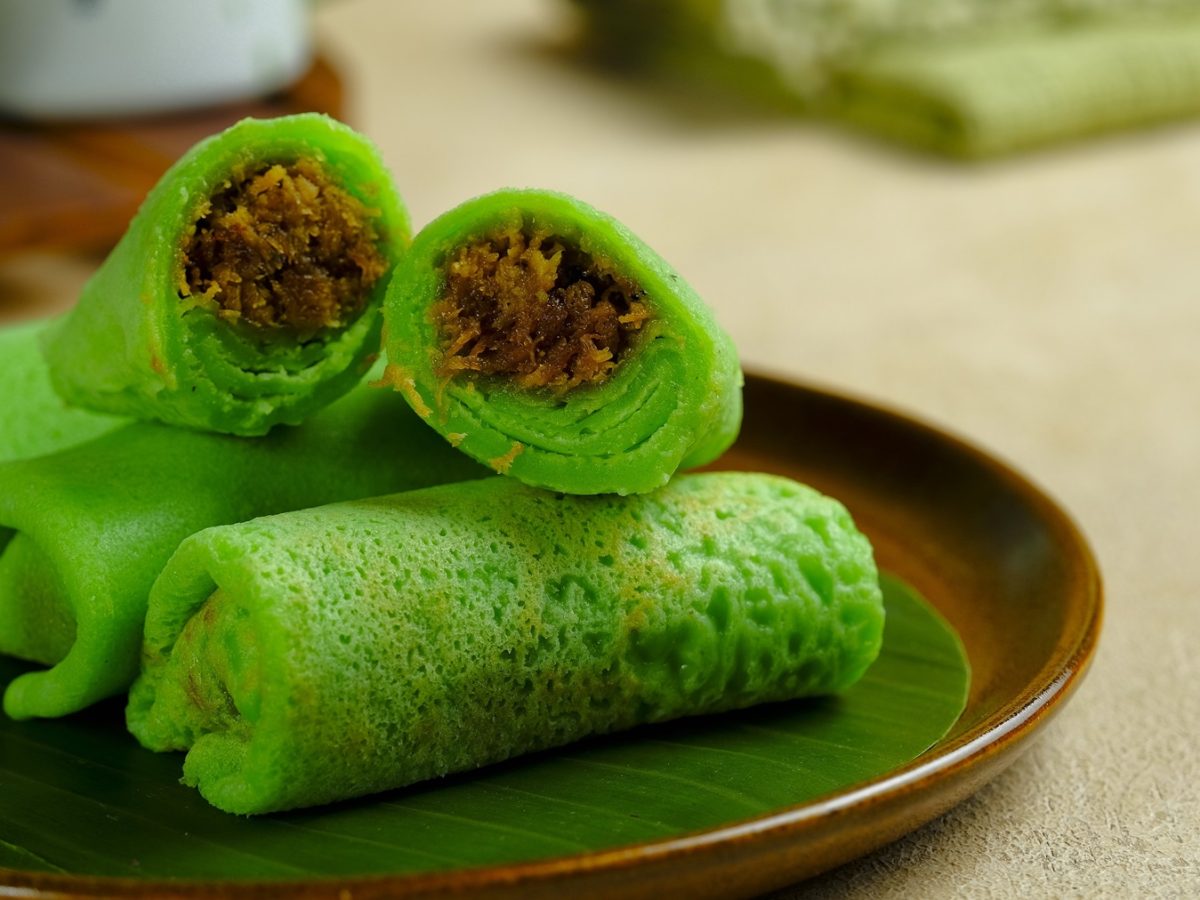 Die grünen Pfannkuchen-Röllchen mit Kokosfüllung auf einem schwarzen Teller und hellem Holztisch. Alles nah und von leicht schräg oben fotografiert.