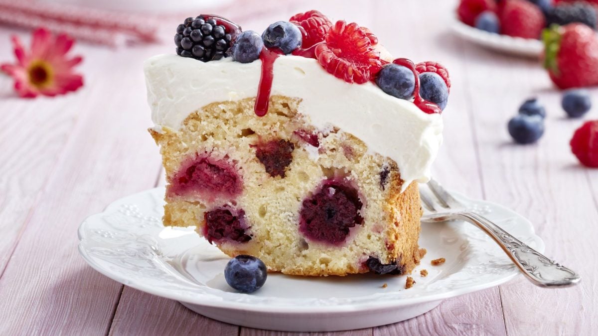 Ein Stück Beeren-Schmand-Kranz auf einem weißen Teller vor rosa Hintergrund. Hinten auf dem Foto Beeren und Blumen verteilt.