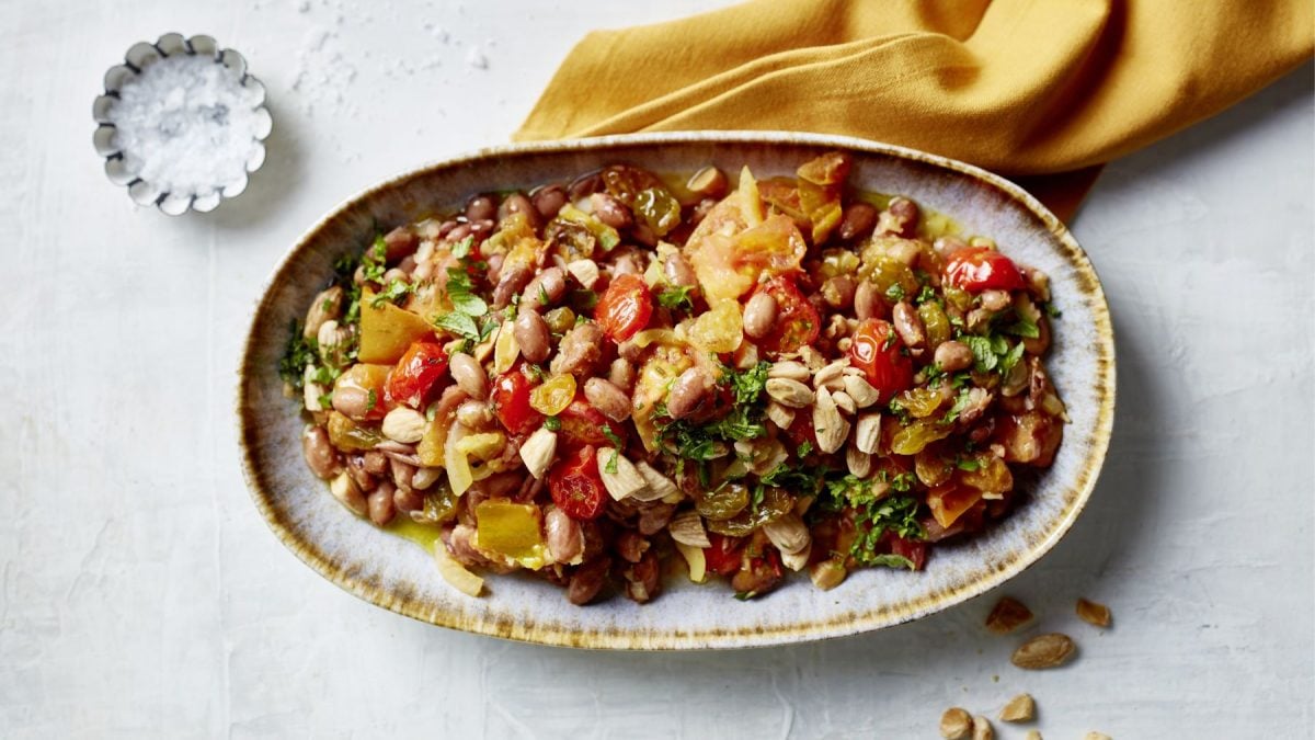 Borlottisalat mit Tomaten und Salzmandeln auf einem grauen Servierteller vor grauem Hintergrund. Darauf ein Schüsselchen mit Salz und ein gelbes Geschirrtuch.