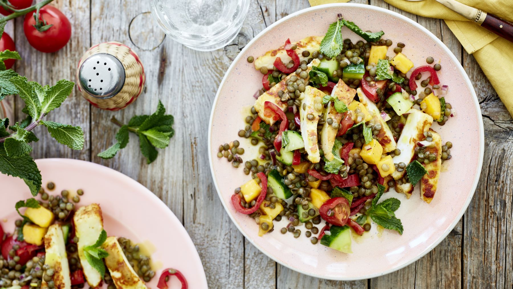 Lauwarmer Linsensalat mit Halloumi auf zwei Tellern verteilt auf hölzernem Hintergrund. Rechts oben auf dem Foto eine gelbe Serviette und links Tomaten, Minze und Pfeffer.